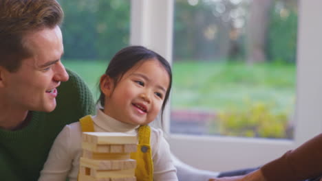 Daughter-Whispering-In-Parent's-Ear-As-Family-With-Two-Dads-Play-Game-Stacking-Wooden-Bricks