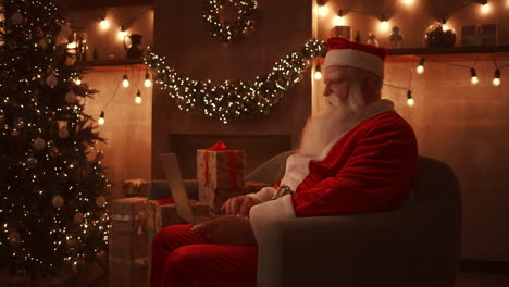Der-Weihnachtsmann-Sitzt-Auf-Einem-Weichen-Sofa-In-Einem-Wunderschön-Dekorierten-Wohnzimmer-Mit-Lichtern-Und-Girlanden.-Er-Hält-Einen-Laptop-Auf-Seinem-Schoß-Und-Tippt-Mit-Einer-Hand-Text.-Hochwertiges-4K-Filmmaterial