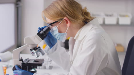 Female-Lab-Research-Worker-In-PPE-Looking-Through-Microscope-And-Recording-Results-On-Laptop