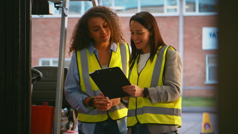 Female-Manager-With-Clipboard-In-Warehouse-With-Female-Worker-Standing-By-Fork-Lift-Truck