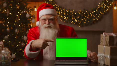 Im-Hintergrund-Des-Weihnachtsbaums-Hält-Ein-älterer-Weihnachtsmann-Einen-Laptop-Und-Zeigt-Mit-Einem-Chromakey-Auf-Den-Bildschirm.-Laptop-Mit-Grünem-Bildschirm.-Der-Weihnachtsmann-Schaut-In-Die-Kamera.-Hochwertiges-4K-Filmmaterial