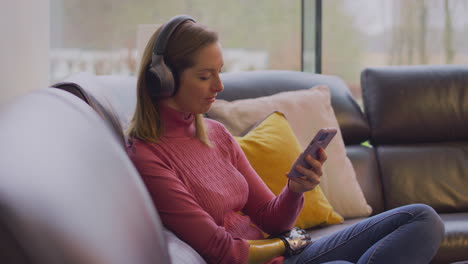 Woman-With-Prosthetic-With-Wireless-Headphones-Listening-To-Music-On-Mobile-Phone-On-Sofa