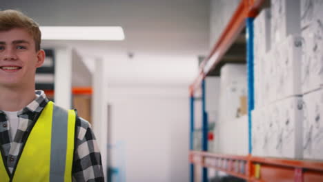 Portrait-Of-Smiling-Young-Male-Worker-Wearing-High-Vis-Safety-Vest-Holding-Box-Inside-Warehouse