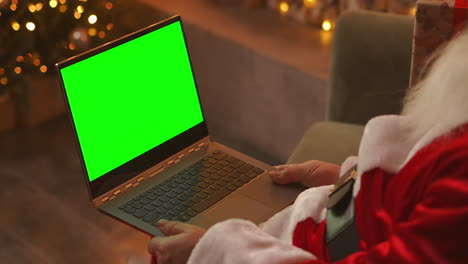 Der-Weihnachtsmann-Führt-Einen-Videoanruf-über-Seinen-Laptop.-Verwenden-Sie-Den-Grünen-Bildschirm-Für-Einen-Videoanruf.-Der-Weihnachtsmann-Grüßt-Mit-Seiner-Hand-Und-Schaut-Auf-Den-Grünen-Laptop-Bildschirm.-Hochwertiges-4K-Filmmaterial