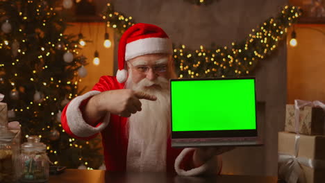 In-the-background-of-the-Christmas-tree-an-elderly-Santa-Claus-is-holding-a-laptop-and-pointing-at-the-screen-with-a-chromakey.-Laptop-with-a-green-screen.-Santa-looks-at-the-camera.-High-quality-4k-footage