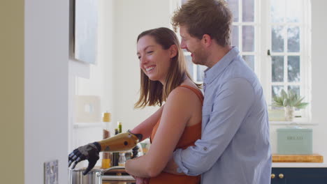 Loving-Couple-With-Woman-With-Prosthetic-Arm-In-Kitchen-Preparing-Meal-Together-And-Hugging