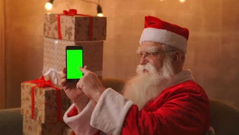 Der-Weihnachtsmann-Sitzt-Auf-Dem-Sofa-Vor-Einem-Weihnachtsbaum-Und-Girlanden-Und-Hält-Ein-Mobiltelefon,-Auf-Das-Ein-Grüner-Bildschirm-Zeigt,-Mit-Dem-Finger.-Der-Weihnachtsmann-Hält-Ein-Telefon-Mit-Einem-Chromakey-Auf-Dem-Bildschirm