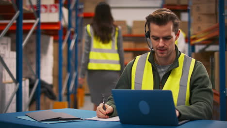 Male-Worker-Wearing-Headset-Sitting-At-Desk-Working-On-Laptop-In-Busy-Warehouse