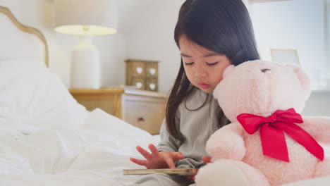 Young-Girl-Sitting-On-Bed-With-Mobile-Phone-Wearing-Pyjamas-At-Home-Cuddling-Soft-Toy