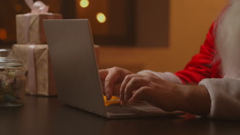 Hands-Santa-typing-on-wireless-keyboard-by-wooden-New-Year-decorated-table-Santa-Claus-is-working-with-a-laptop-looking-through-mail-and-answering-messages-to-children.-High-quality-4k-footage