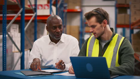 Manager-With-Male-Worker-Wearing-Headset-Sitting-At-Desk-Working-On-Laptop-In-Busy-Warehouse