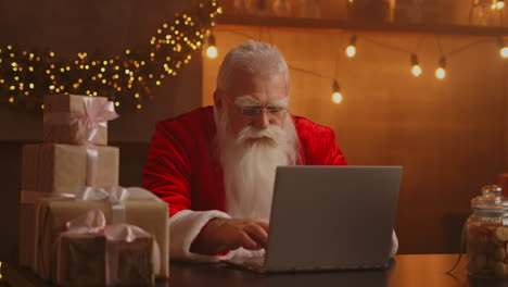 Real-Santa-Claus-using-new-technology-for-communication-with-children-receiving-mail-or-wish-list.-Cheerful-working-on-laptop-and-smiling-while-sitting-at-his-chair-with-fireplace-and-Christmas-Tree.-High-quality-4k-footage