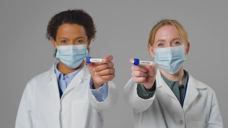 Studio-Shot-Of-Lab-Research-Workers-In-Face-Masks-Holding-Test-Tubes-Labelled-Covid-19-And-Vaccine