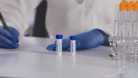 Close-Up-Of-Female-Lab-Research-Worker-In-PPE-Studying-Test-Tubes-Labelled-Omicron-And-Vaccine