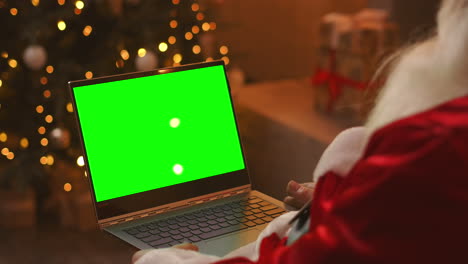 Der-Weihnachtsmann-Führt-Einen-Videoanruf-über-Seinen-Laptop.-Verwenden-Sie-Den-Grünen-Bildschirm-Für-Einen-Videoanruf.-Der-Weihnachtsmann-Grüßt-Mit-Seiner-Hand-Und-Schaut-Auf-Den-Grünen-Laptop-Bildschirm.-Hochwertiges-4K-Filmmaterial