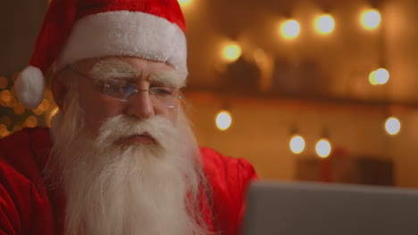 Weihnachtsmann-Benutzt-Laptop-Im-Wohnzimmer.-Porträt-Des-Nachdenklichen-Weihnachtsmanns,-Der-Mit-Modernem-Laptop-In-Dekoriertem-Haus-Tippt.-Hochwertiges-4K-Filmmaterial