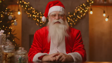 Santa-Claus-is-sitting-at-a-table-in-a-beautiful-living-room-decorated-for-a-merry-christmas.-He-waves-his-right-hand-and-starts-talking.-High-quality-4k-footage