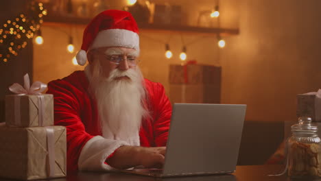 Santa-Claus-holding-gift-box-typing-keyboard-kid-talking-to-child-greeting-on-Merry-Christmas-Happy-New-Year-in-virtual-online-chat-on-laptop-sitting-at-home-table-late-with-present-on-xmas-eve.-High-quality-4k-footage