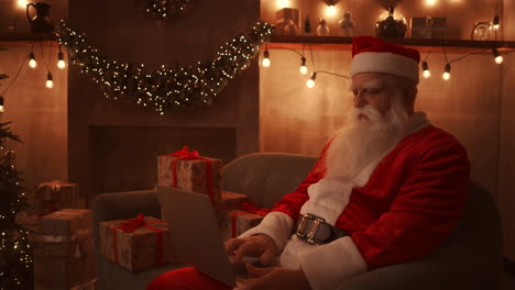 Der-Weihnachtsmann-Sitzt-Auf-Einem-Bequemen-Weichen-Sofa,-Hält-Einen-Laptop-Auf-Seinem-Schoß-Und-Tippt-Darauf.-Schönes-Wohnzimmer,-Dekoriert-Für-Ein-Treffen,-Frohe-Weihnachten-Und-Ein-Gutes-Neues-Jahr.-Hochwertiges-4K-Filmmaterial