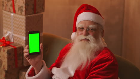 Der-Weihnachtsmann-Sitzt-Auf-Dem-Sofa-Vor-Einem-Weihnachtsbaum-Und-Girlanden-Und-Hält-Ein-Mobiltelefon,-Auf-Das-Ein-Grüner-Bildschirm-Zeigt,-Mit-Dem-Finger.-Der-Weihnachtsmann-Hält-Ein-Telefon-Mit-Einem-Chromakey-Auf-Dem-Bildschirm