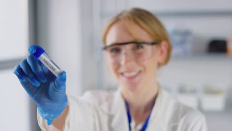 Lächelnde-Labormitarbeiterin-Mit-Schutzbrille-Und-Reagenzglas-Mit-Der-Aufschrift-„Impfstoff“.