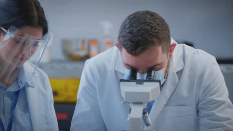 Male-And-Female-Lab-Workers-Conducting-Research-Using-Microscope-And-Writing-Down-Test-Results