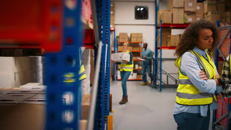 Female-Team-Leader-With-Digital-Tablet-In-Busy-Warehouse-Training-Male-Intern-Standing-By-Shelves