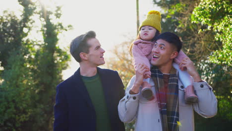 Family-With-Two-Dads-On-Walk-In-Winter-Countryside-Carrying-Daughter-On-Shoulders