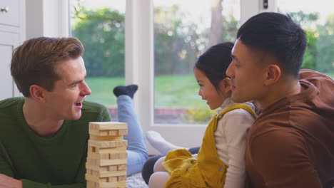 Daughter-Whispering-In-Parent's-Ear-As-Family-With-Two-Dads-Play-Game-Stacking-Wooden-Bricks
