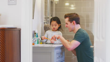 Family-With-Dad-Helping-Daughter-With-Toothpaste-As-She-Brushes-Teeth-In-Bathroom