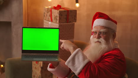 Der-Weihnachtsmann-Hält-Einen-Laptop-Mit-Einem-Grünen-Bildschirm,-Während-Er-In-Der-Weihnachtsdekoration-Sitzt.-Hochwertiges-4K-Filmmaterial