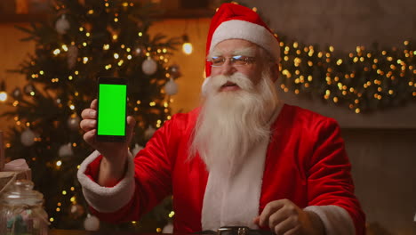 Santa-Claus-is-sitting-on-the-sofa-in-the-background-of-a-Christmas-tree-and-garlands-holding-a-mobile-phone-with-a-green-screen-pointing-at-it-with-his-finger.-Santa-holds-a-phone-with-a-chromakey-on-the-screen