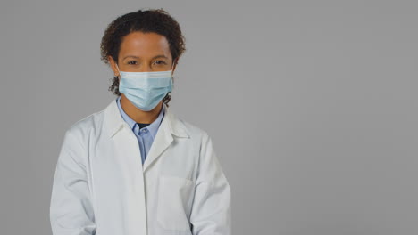 Studio-Portrait-Of-Smiling-Female-Doctor-Or-Lab-Worker-Wearing-Face-Mask-In-White-Coat
