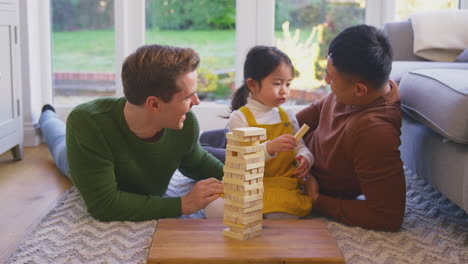 Family-With-Two-Dads-Playing-Game-With-Daughter-At-Home-Stacking-Wooden-Bricks-Into-Tower