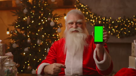 Kind-Santa-Claus-is-holding-a-smartphone-with-a-green-screen.-Indicates-the-smartphone-screen.-Sits-in-a-beautiful-room-decorated-for-a-merry-christmas.-High-quality-4k-footage