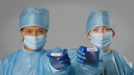 Studio-Shot-Of-Female-Lab-Research-Workers-In-PPE-Holding-Test-Tubes-Labelled-Covid-19-And-Vaccine