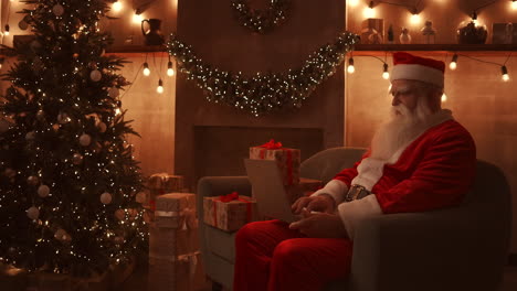 Der-Weihnachtsmann-Sitzt-Auf-Einem-Bequemen,-Weichen-Sofa,-Hält-Einen-Laptop-Auf-Dem-Schoß-Und-Tippt-Darauf.-Hochwertiges-4K-Filmmaterial