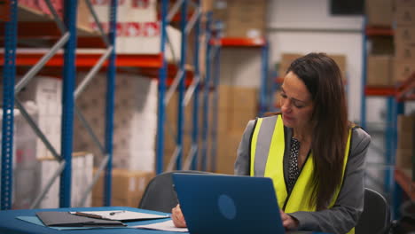 Female-Manager-Wearing-High-Vis-Vest-Sitting-At-Desk-Working-On-Laptop-In-Busy-Warehouse