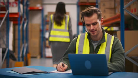 Male-Worker-Wearing-Headset-Sitting-At-Desk-Working-On-Laptop-In-Busy-Warehouse