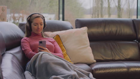 Woman-Wearing-Wireless-Headphones-Listening-To-Music-Streaming-From-Mobile-Phone-On-Sofa-At-Home
