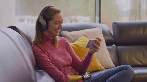 Woman-With-Prosthetic-Arm-Wearing-Wireless-Headphones-Listening-To-Music-On-Mobile-Phone-On-Sofa