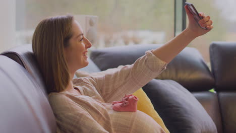 Pregnant-Woman-Sitting-On-Sofa-At-Home-With-Small-Baby-Shoes-On-Stomach-Taking-Selfie