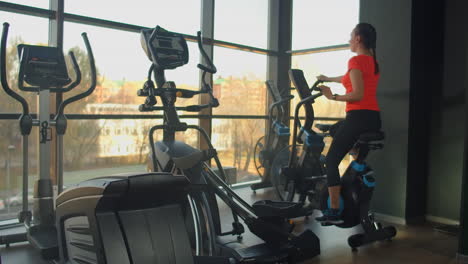 Young-active-woman-spinning-a-air-bike-in-gym-with-trainers.-Female-training-on-air-bike