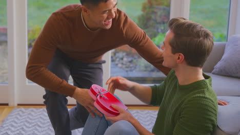Man-Surprising-Same-Sex-Partner-At-Home-With-Heart-Shaped-Gift-Held-Behind-His-Back