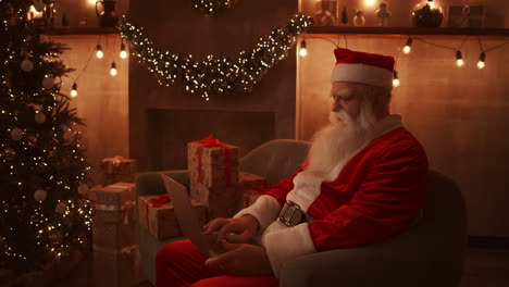 Der-Weihnachtsmann-Sitzt-Neben-Einem-Weihnachtsbaum-Auf-Einem-Bequemen,-Weichen-Sofa,-Hält-Einen-Laptop-Auf-Dem-Schoß-Und-Beantwortet-E-Mails-Von-Kleinen-Kindern.-Hochwertiges-4K-Filmmaterial
