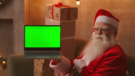 In-the-background-of-the-Christmas-tree-an-elderly-Santa-Claus-is-holding-a-laptop-and-pointing-at-the-screen-with-a-chromakey.-Laptop-with-a-green-screen.-Santa-looks-at-the-camera.-High-quality-4k-footage