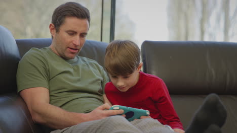 Father-with-son-wearing-pyjamas-sitting-on-sofa-at-home-playing-computer-game-on-handheld-device---shot-in-slow-motion