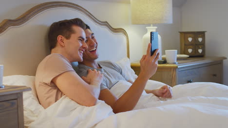 Gleichgeschlechtliches-Männliches-Paar,-Das-Zu-Hause-Im-Bett-Liegt-Und-Einen-Videoanruf-Auf-Dem-Mobiltelefon-Tätigt