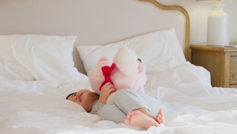 Young-Girl-Playing-On-Bed-Wearing-Pyjamas-At-Home-Cuddling-Soft-Toy