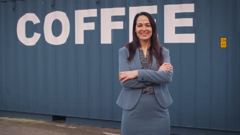 Portrait-Of-Female-Freight-Haulage-Manager-Standing-By-Shipping-Container-For-Coffee